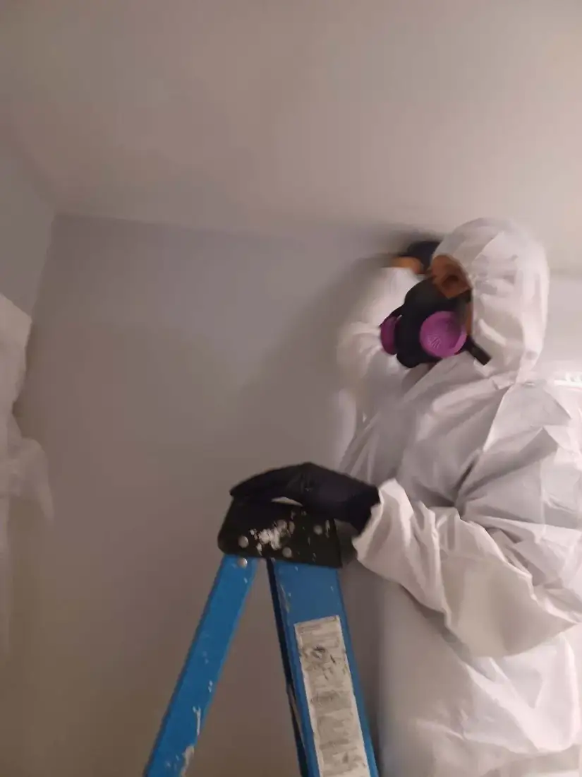 Technician in protective gear and a respirator repairing a ceiling from a ladder