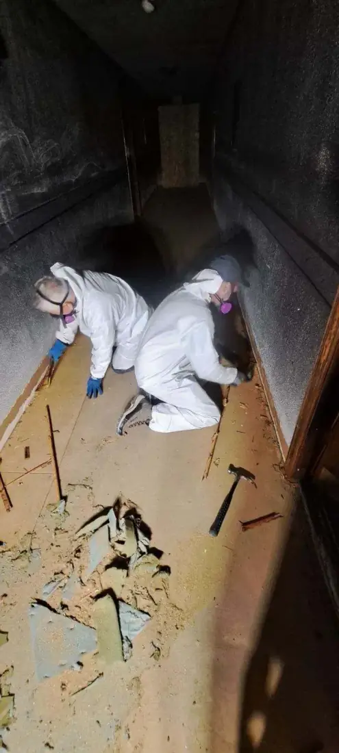 Two technicians in protective gear demolishing a dark, fire damaged corridor