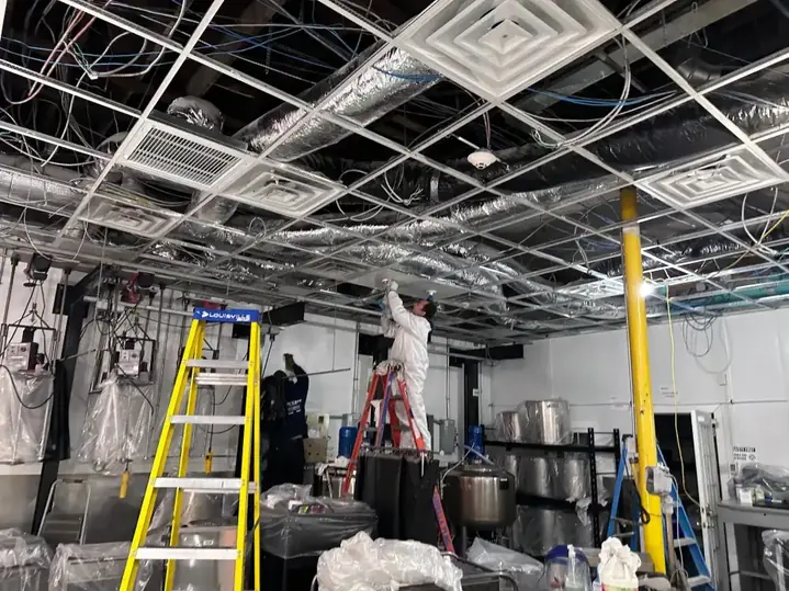 Technician on a ladder working above exposed ductwork and ceiling grid in a commercial space